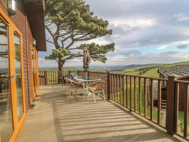 A wooden deck with a glass table chairs an umbrella and a tree overlooking green hills at Lodge 23 in Torpoint