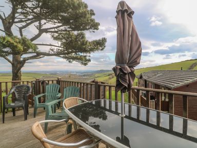 An outdoor deck with a glass table, several chairs, a closed umbrella, wooden railing, and a view of green hills and cabins at Lodge 23 in Torpoint