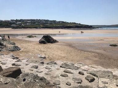 A sandy beach with large rocks and people walking near water with houses on a hill in the background at Lowenda in Polzeath