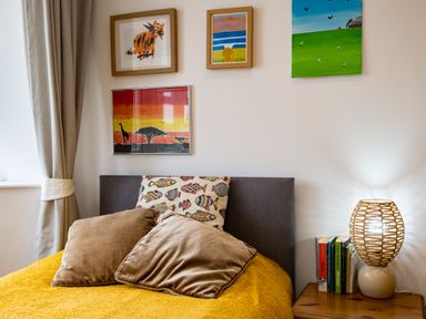 A bedroom with a bed with beige pillows and a fish-patterned cushion a wooden bedside table with books and a lamp and four framed paintings on the wall at Lowenda in Polzeath