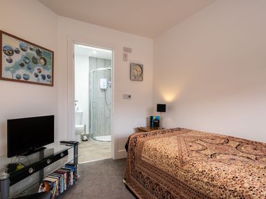 A bedroom with a bed a bedside table with a lamp a wall clock a tv on a glass stand with books and a view into an en suite bathroom at Lowenda in Polzeath
