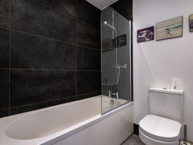 A bathroom with a bathtub featuring a glass shower screen and a toilet with picture frames on the wall at Lowenda in Polzeath