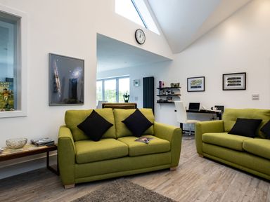 A living room with two green sofas and black pillows a small wooden side table two framed pictures a wall clock and a white chair with a black desk at Lowenda in Polzeath