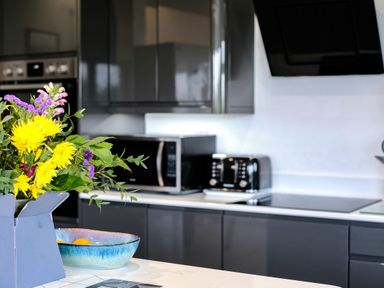 A modern kitchen with a bouquet of flowers on the counter a microwave and toaster on the countertop and dark cabinets at Lowenda in Polzeath