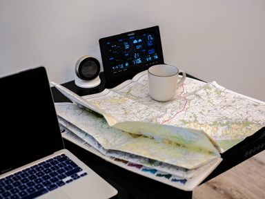 A desk with a laptop a folded map a white mug and two digital devices a smart thermostat and a weather station