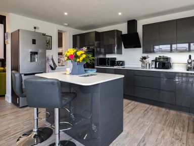 A kitchen with grey cabinets a kitchen island with two black bar stools and flowers in a vase on the island at Lowenda in Polzeath