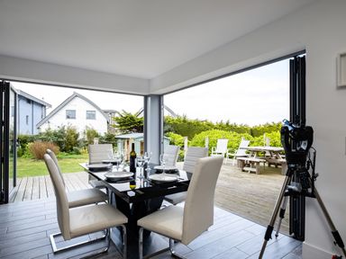 A dining area with six chairs around a table set with plates and glasses next to an open door leading to an outdoor deck with picnic table and chairs at Lowenda in Polzeath