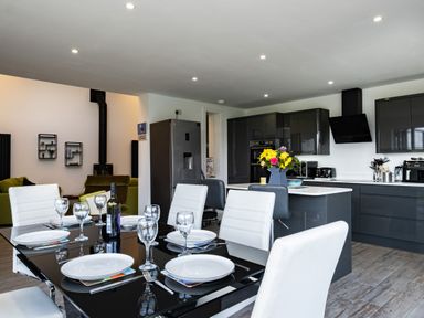 A modern kitchen and dining area with a black glass table set for six and a kitchen island with flower vase at Lowenda in Polzeath