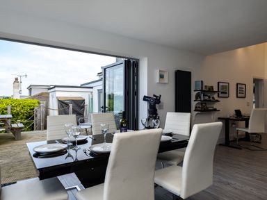 A dining area with a black table set with plates and glasses next to an open glass door leading to an outdoor patio at Lowenda in Polzeath