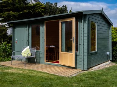 A green garden shed with an open wooden door and windows a metal bench with pillows outside on a wooden deck at Lowenda in Polzeath