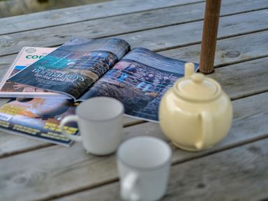 A wooden table with a teapot two mugs and magazines including an open magazine showing a photo of people on a bridge at Lowenda in Polzeath