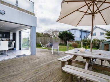 An outdoor wooden deck with a picnic table and umbrella adjacent to a dining area with white chairs in a house at Lowenda in Polzeath