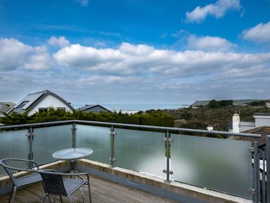 A balcony with two chairs and a round table overlooking houses trees and the sea at Lowenda in Polzeath