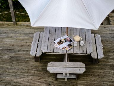 A wooden picnic table with a teapot two mugs and magazines under a white patio umbrella on a wooden deck at Lowenda in Polzeath