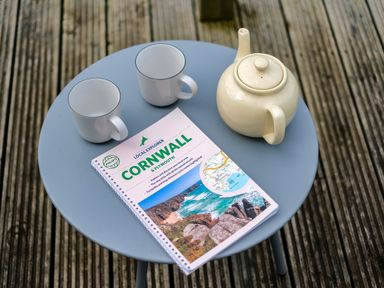 A round blue table with two white mugs a white teapot and a Cornwall guidebook on a wooden deck at Lowenda in Polzeath
