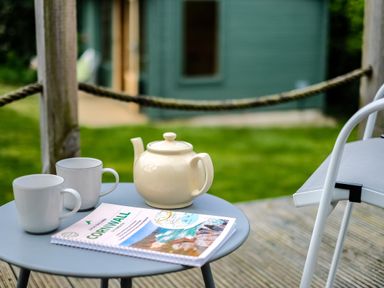 A round table with two white mugs a teapot and a Cornwall travel guide on a deck outside a green building at Lowenda in Polzeath