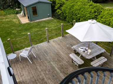 A wooden deck with a picnic table and white umbrella next to a grass lawn and green shed at Lowenda in Polzeath