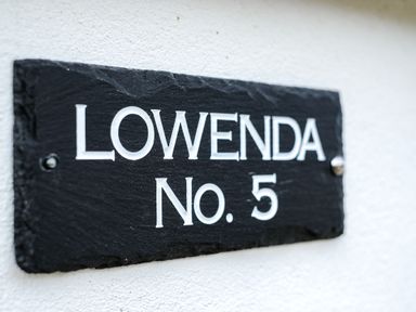 A black slate house nameplate with white text reading Lowenda No 5 on a white wall at Lowenda Polzeath