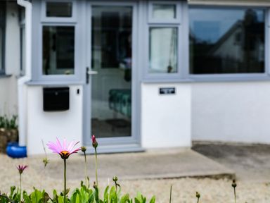 A flower in front of a grey door and white wall with a window at Lowenda in Polzeath