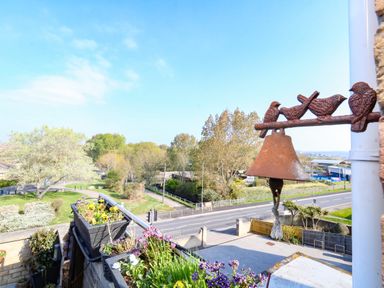 A balcony with flower pots and a metal bird bell overlooking a road and green trees at Beach Retreat in Weymouth