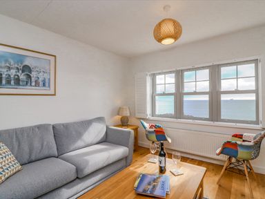 A living room with a grey sofa two colorful chairs a wooden table with wine glasses and books and a window with a sea view at Beach Retreat in Weymouth