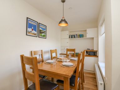 A dining room with a wooden table set with plates and glasses six chairs a pendant light built-in shelves with books and a window at Beach Retreat in Weymouth
