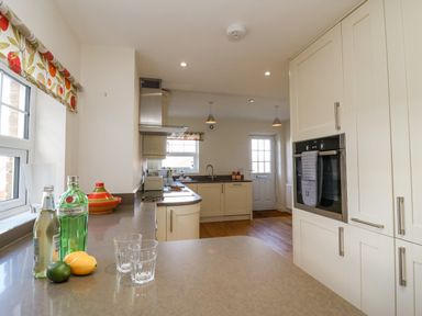 A kitchen with white cabinets, a built-in oven, a countertop with bottles and glasses, and a window with a patterned blind at Beach Retreat in Weymouth