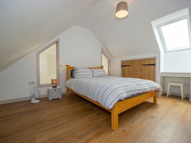 A bedroom with a wooden bed frame, striped bedding, wooden floor, side table with lamp and fan, built-in cupboard, and skylight window at Owl Lodge Wedmore