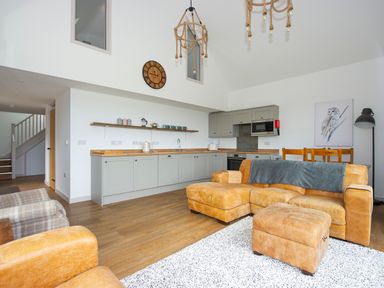 An open plan living room and kitchen with tan leather sofa and ottoman wooden dining chairs and gray kitchen cabinets at Owl Lodge in Wedmore