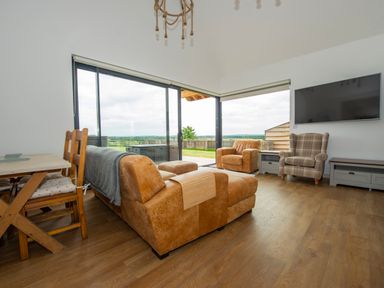 A living room with a brown sofa set, wooden dining table with chairs, large windows, and a wall mounted TV at Owl Lodge in Wedmore