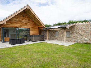 An outdoor area with grass a wooden building a hot tub and a stone wall with a covered patio at Owl Lodge in Wedmore