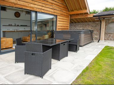 A patio with black wicker table and chairs near a hot tub outside a wooden building at Owl Lodge in Wedmore