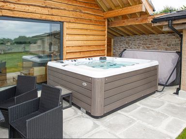 An outdoor patio with a hot tub black wicker chairs and wooden and stone walls at Owl Lodge in Wedmore