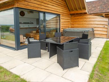 An outdoor patio with a black dining table and chairs next to a wooden building with sliding glass doors and a hot tub at Starling Lodge in Wedmore