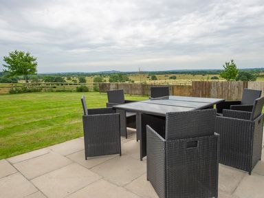 An outdoor patio with wicker chairs and a glass table overlooking a grassy field and distant trees at Starling Lodge in Wedmore