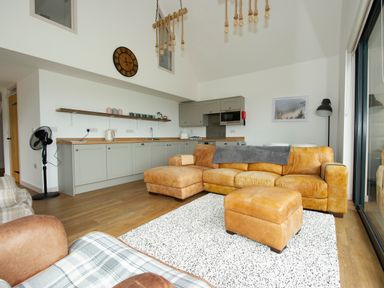 A living room with a tan leather sofa a matching ottoman a white patterned rug and a kitchen with gray cabinets at Starling Lodge in Wedmore