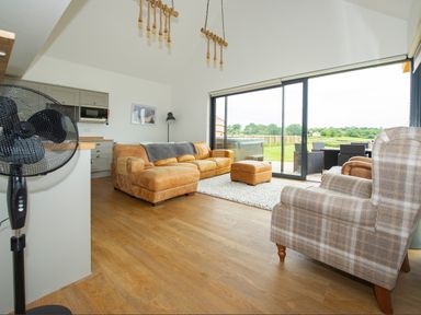 A living room with brown leather sofa and plaid armchair near large sliding glass doors showing outdoor patio at Starling Lodge in Wedmore