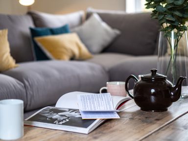 A wooden coffee table with a teapot mugs open books and a vase with green leaves in front of a sofa with cushions at Samphire in Rock