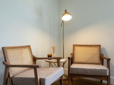 Two wooden armchairs with woven backs a small round table with a candle and reed diffuser and a floor lamp in a corner Samphire Rock