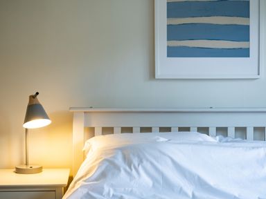 A bedroom with a white bed frame, white bedding, a bedside table with a lamp, and a blue abstract painting on the wall at Samphire in Rock
