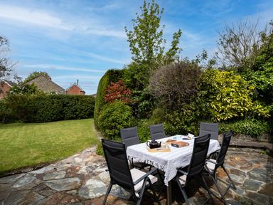 An outdoor dining area with a table covered by a white cloth and six chairs on a stone patio surrounded by green bushes and trees at Samphire in Rock
