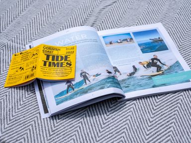 A booklet titled Tide Times 2022 on a magazine page showing people surfing on water at Samphire in Rock