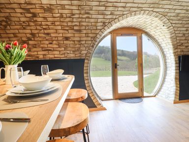 A dining area with a wooden table set with plates and glasses next to a round brick wall with a circular door showing an outdoor view at Nant y Garnedd in Mallwyd near Dinas Mawddwy