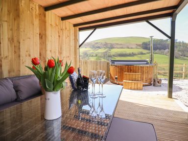 A covered patio with a glass table holding a vase of red tulips and champagne glasses next to a hot tub with wooden steps and a view of green hills at Nant y Garnedd in Mallwyd near Dinas Mawddwy