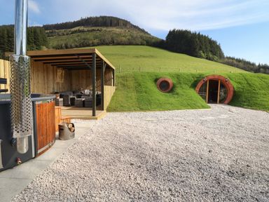 An outdoor area with a hot tub and covered seating next to a grass-covered hobbit-style house with circular windows and door at Nant yr Esgair Mallwyd near Dinas Mawddwy