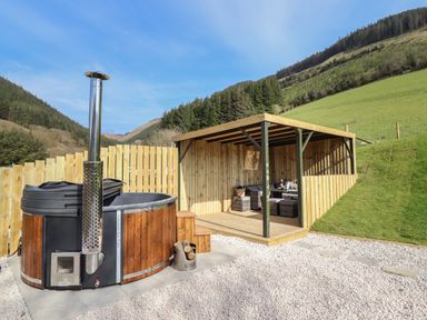 An outdoor hot tub next to a wooden covered seating area in a fenced yard with grassy hills and trees at Nant yr Esgair in Mallwyd near Dinas Mawddwy