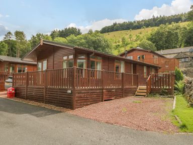 A wooden lodge with a fenced porch surrounded by trees and hills at Kirkstone Lodge in Limefitt Park near Windermere