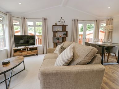 A living room with a beige sofa, wooden coffee table, shelves, TV on a wooden stand, dining table with chairs and wine glasses at Kirkstone Lodge in Limefitt Park near Windermere