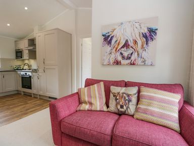 A red sofa with three cushions and a painting of a cow above in a living room next to a kitchen at Kirkstone Lodge Limefitt Park near Windermere