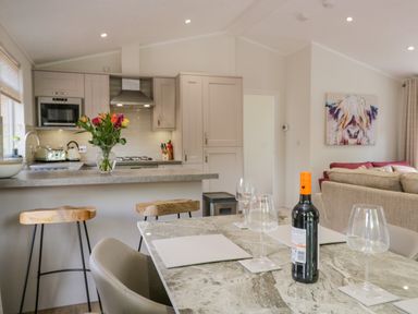 A kitchen with a countertop, stools, a dining table with wine glasses and a bottle, and a living room with a couch and animal artwork at Kirkstone Lodge Limefitt Park near Windermere